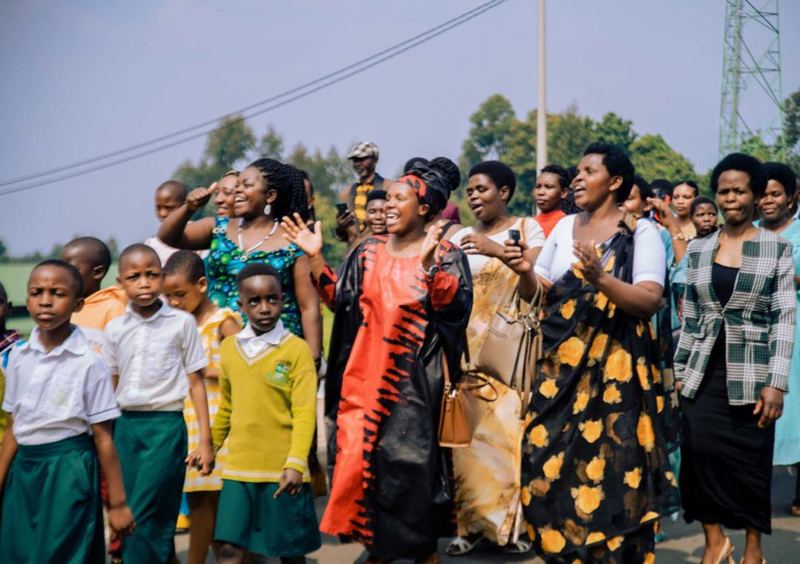 Women in bright dresses clapping as they walk alongside pupils in uniform