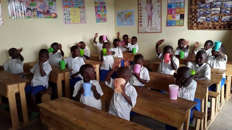 Pupils in white shirts at wooden desks in a classroom with posters on the walls, drinking from coloured plastic cups