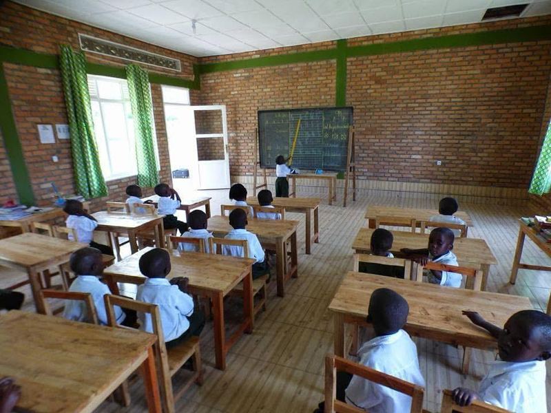 Pupils in white shirts sitting at wooden desks in a brick classroom, one pupil standing at the blackboard