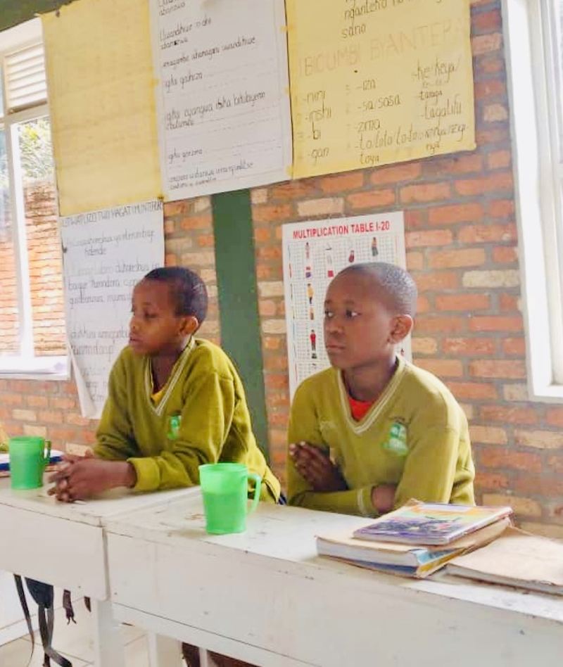 Two pupils in green school jumpers sitting at white desks with green cups and exercise books, handwritten lesson posters on the brick wall behind