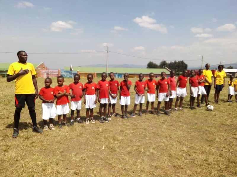 A football team in red shirts and white shorts lined up on a grass field, an adult in a yellow shirt standing in front