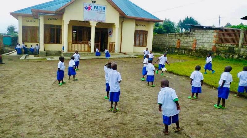Young pupils in white shirts and blue shorts kicking a football on the dirt yard in front of the Faith Foundation building