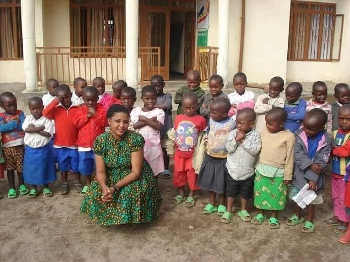Faith Uwantege, the founder, crouching in a green patterned dress in front of a large group of young children with folded arms outside the school