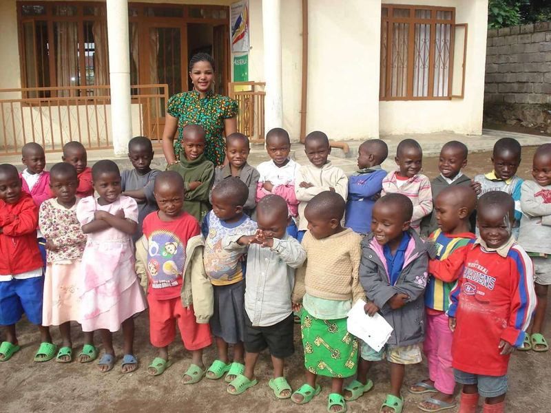 Faith Uwantege, the founder, standing behind a large group of smiling young children with folded arms outside the school