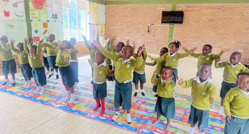 Young pupils in green uniform dancing with their arms raised on a colourful mat in a classroom
