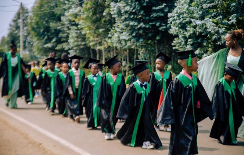 Children in black graduation gowns with green stoles and mortarboards walking along a road