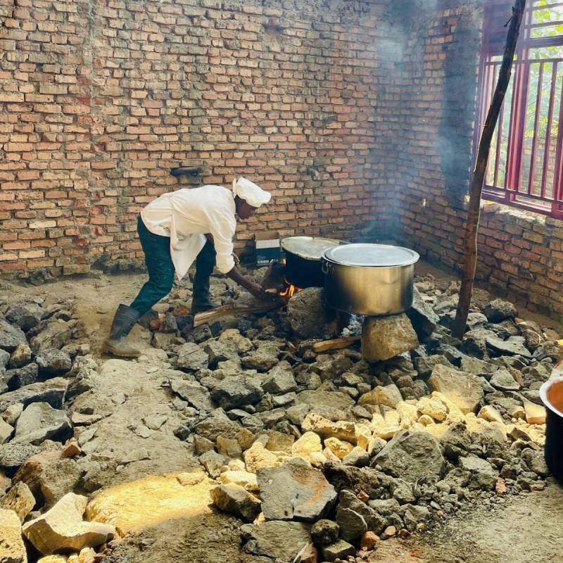 A cook in white tending a wood fire under large cooking pots in a brick-walled outdoor kitchen