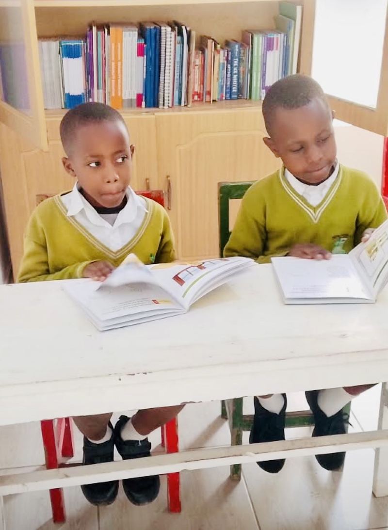 Two pupils in green school jumpers reading picture books at a white table, a bookcase full of books behind them