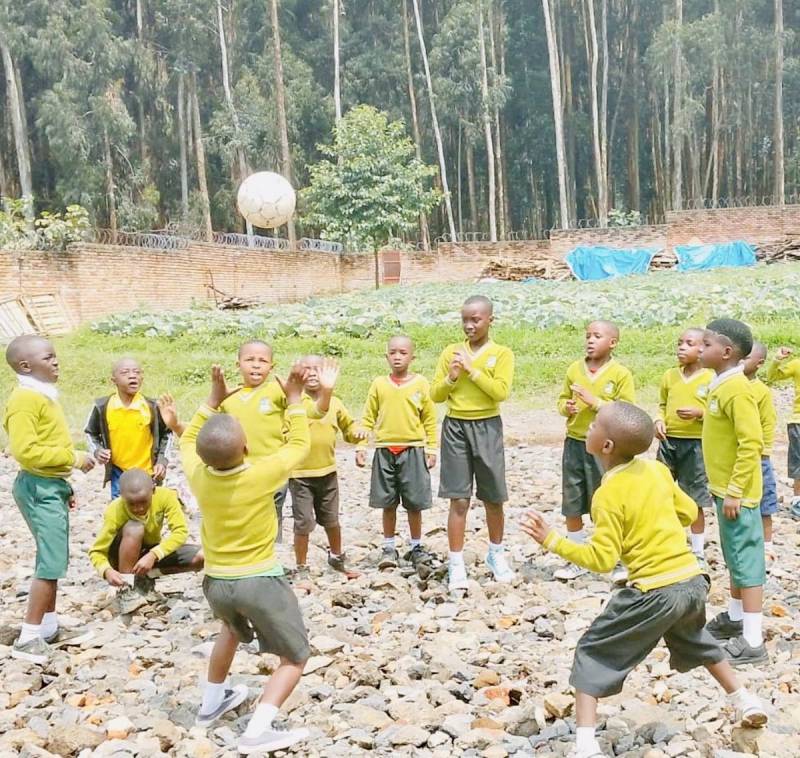 Pupils in yellow school jumpers playing with a ball on open ground in front of tall trees