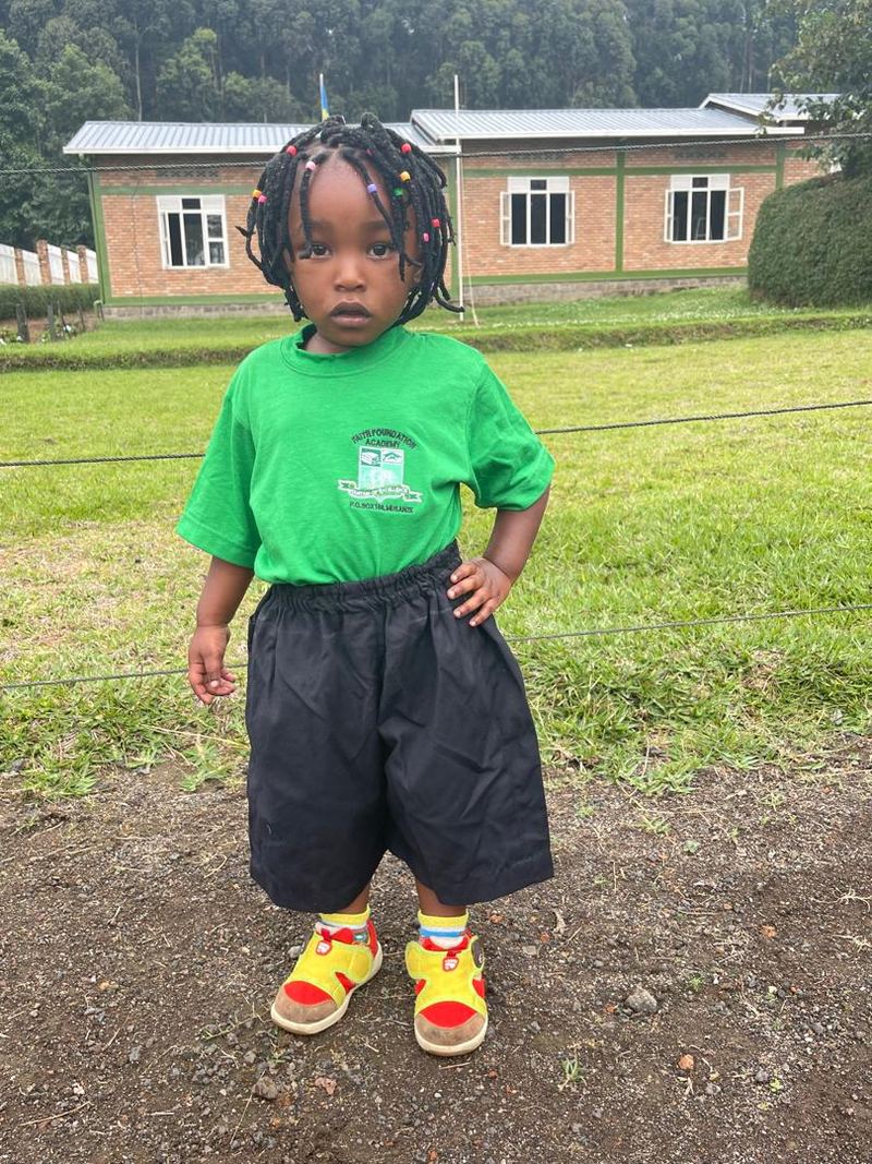 A small girl with braided hair in a green school T-shirt and black shorts standing on the school grounds