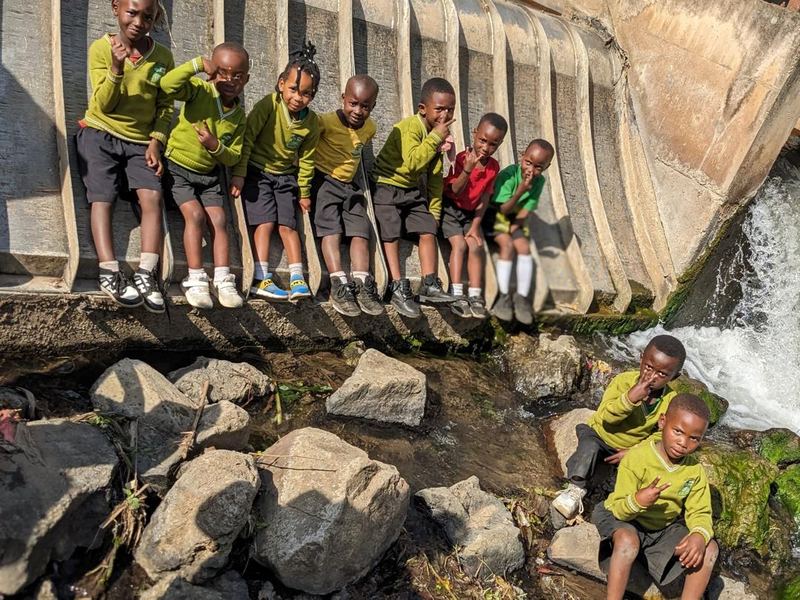 Pupils in yellow jumpers lined up along the edge of a concrete water channel, water pouring down beside them